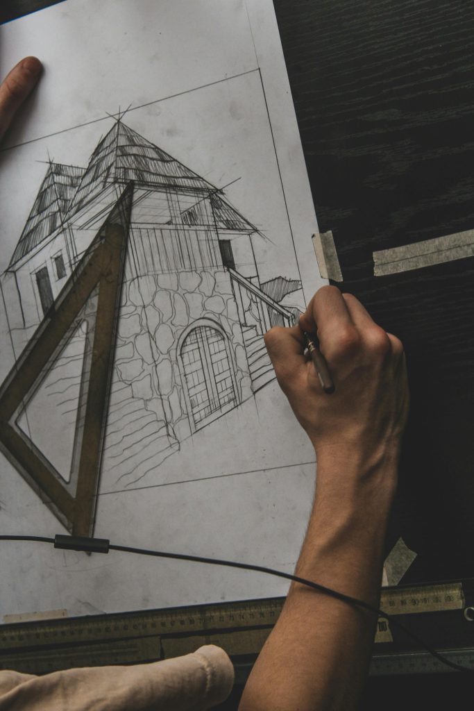 A close-up of an adult hand drawing a house with architectural tools on paper.